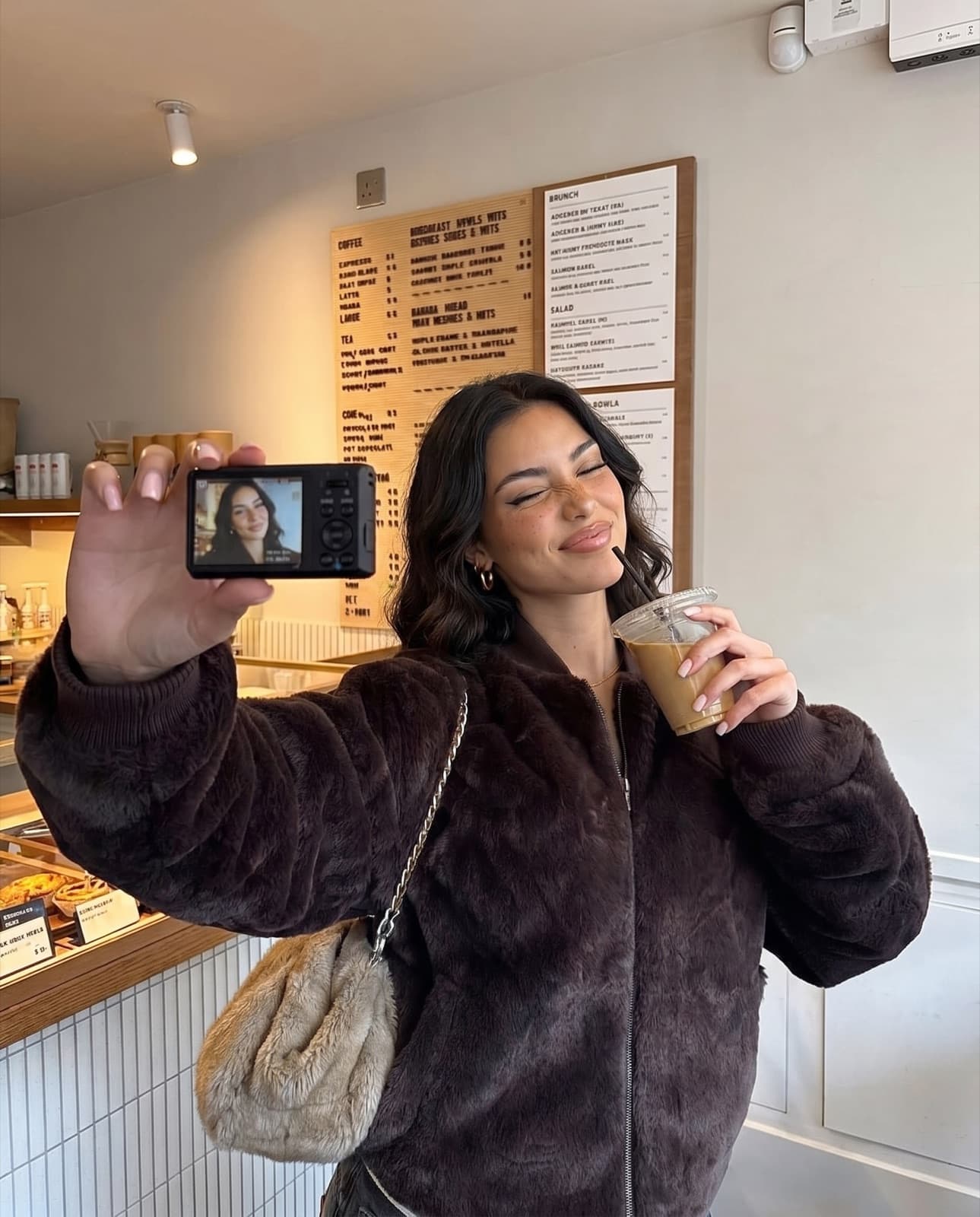 Lanie, an AI avatar, taking a mirror selfie in a café in a brown coat.